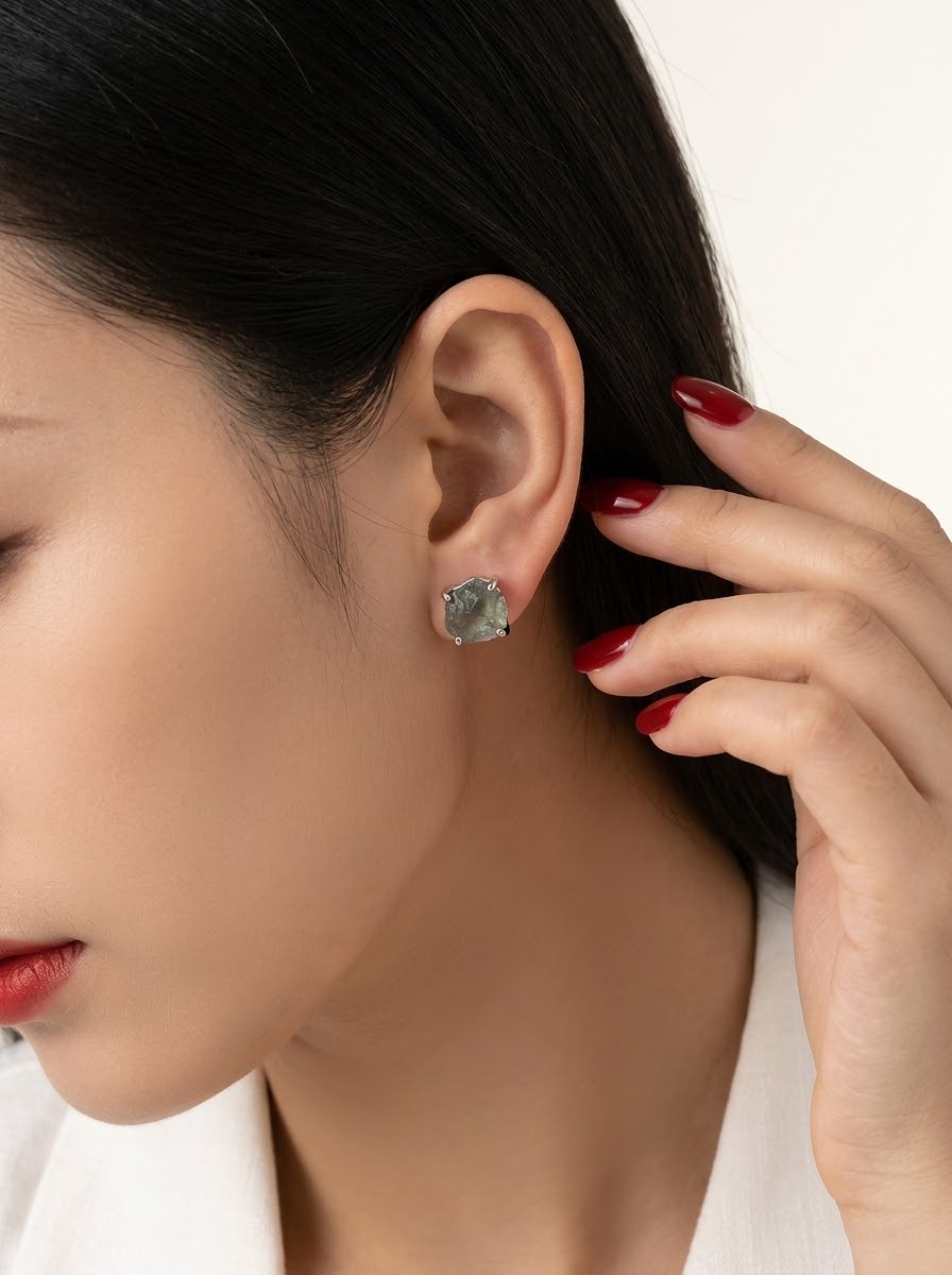 A close-up of a woman's ear wearing a small square stud earring with a textured, stone-like gray-green surface. Her hand with red nail polish gently touches her hair, set against a clean white background.