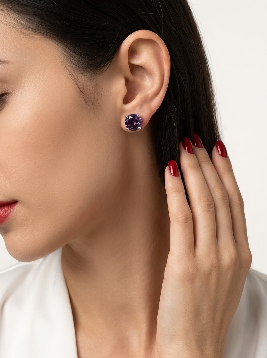 A close-up of a woman wearing a small round stud earring with a deep purple gemstone in a silver-toned setting, with her hand resting near her ear against a clean white background.