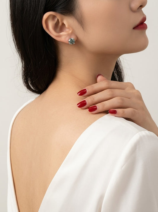 A close-up lifestyle shot of a model wearing a small, round turquoise-blue gemstone stud earring. The model is shown from the back and side, wearing a white off-the-shoulder top, with dark hair, red lipstick, and red manicured nails visible against a clean, light gray background.