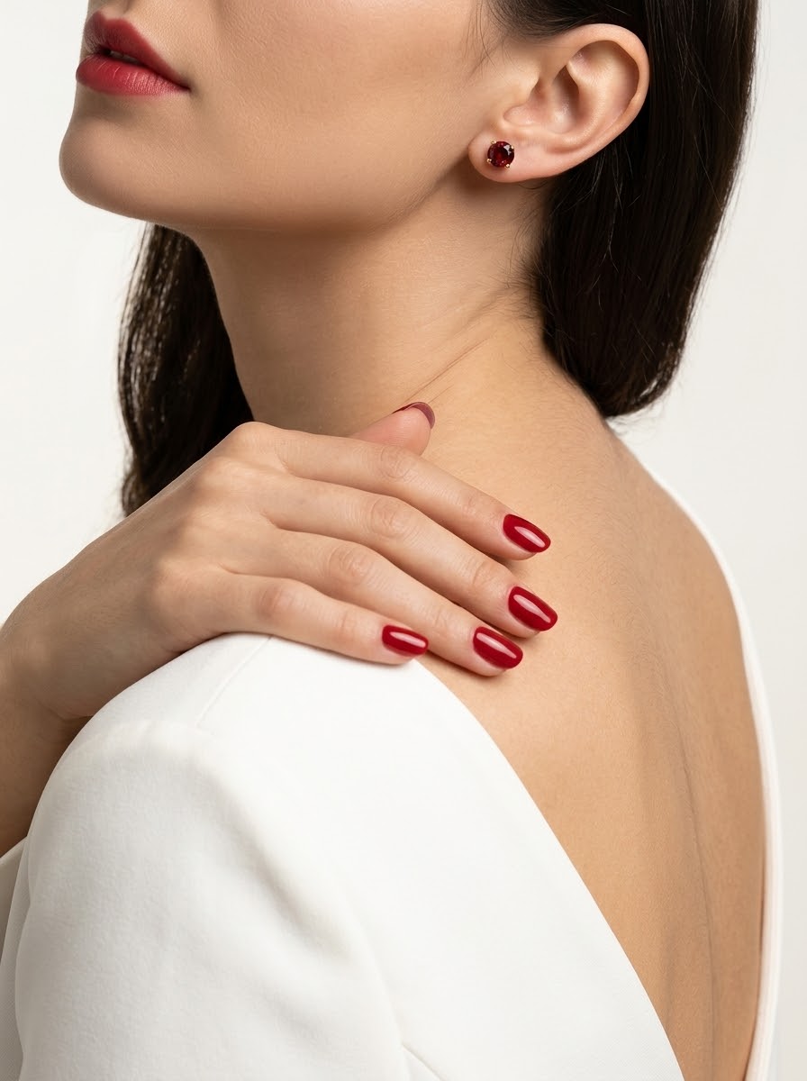 A close-up lifestyle shot of a model wearing a small round red gemstone stud earring, with her hand resting on her shoulder. She is wearing a white top, and her nails are painted a matching deep red.