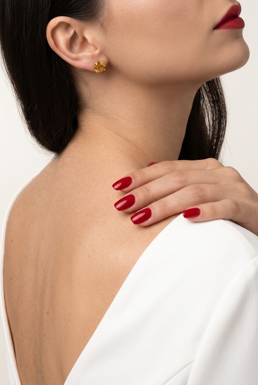 A close-up lifestyle shot of a model wearing a small round yellow gemstone stud earring, with her hand resting on her shoulder and her red manicured nails visible against a white top.