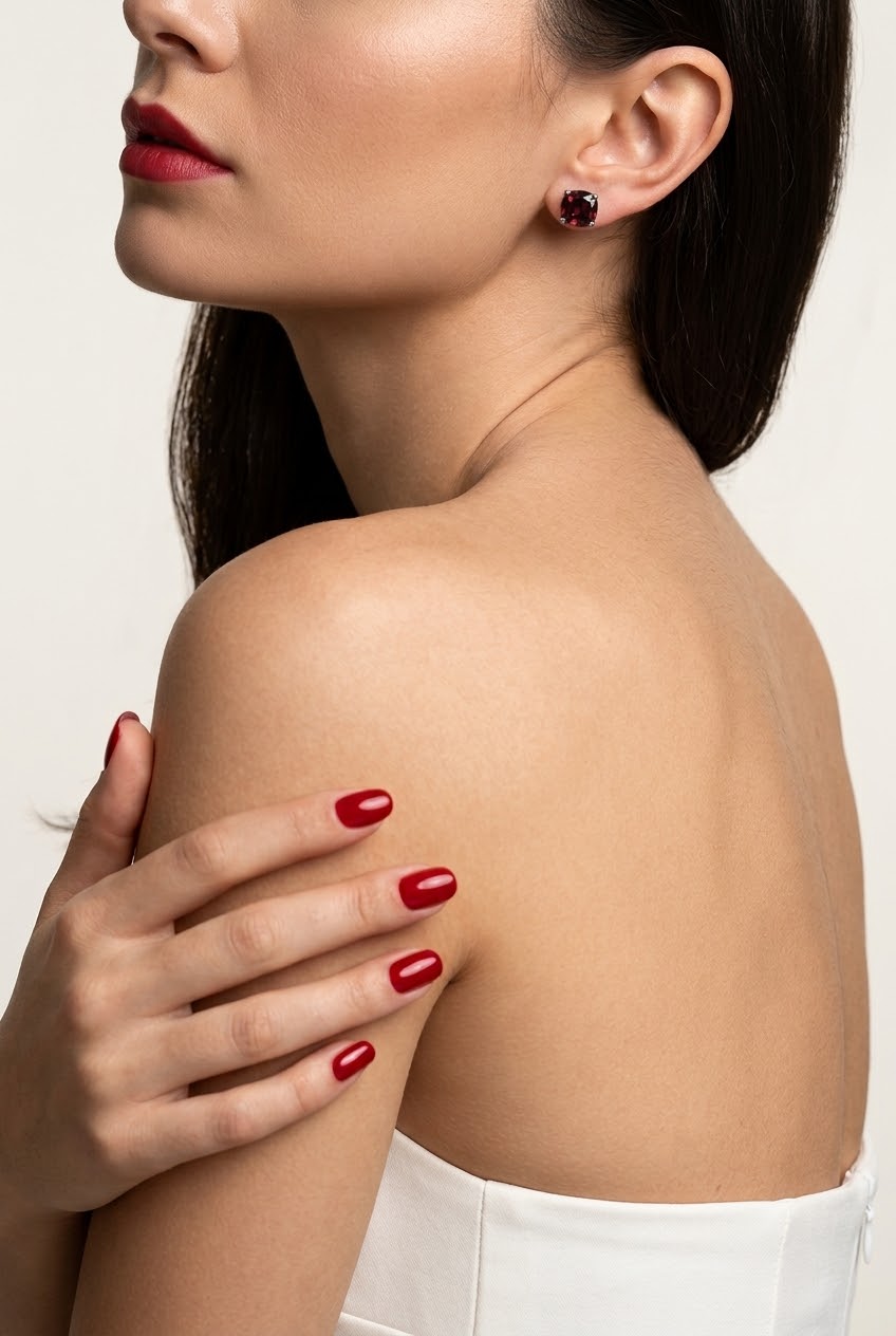 Close-up of a woman wearing a small round dark red stud earring, with her hand resting on her shoulder. She has dark hair, red lipstick, and red nail polish, wearing a white strapless top against a light off-white background.