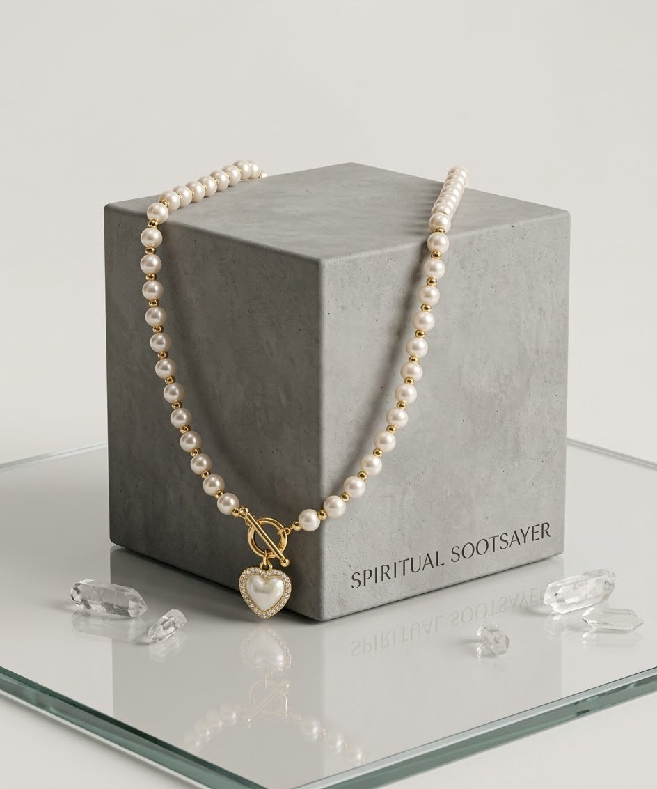 A pearl necklace with a gold toggle clasp and heart-shaped pendant, draped over a gray display box on a glass surface with clear crystal props.