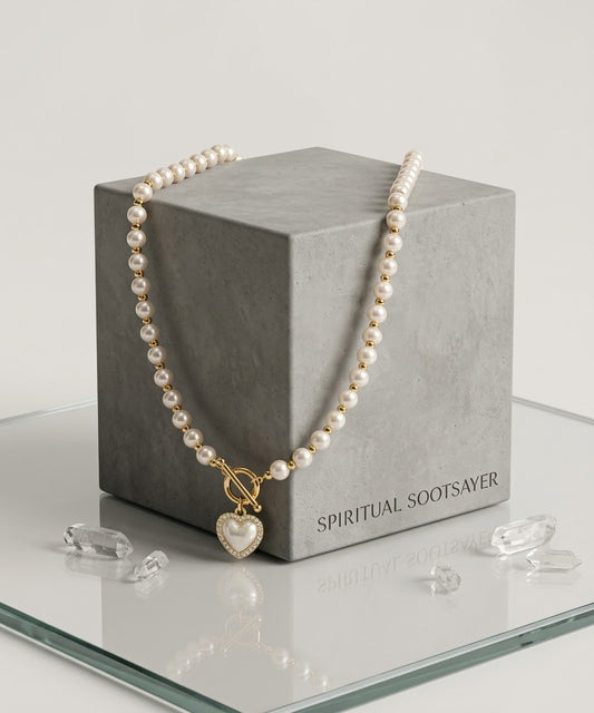 A pearl necklace with a gold toggle clasp and heart-shaped pendant, draped over a gray display box on a glass surface with clear crystal props.