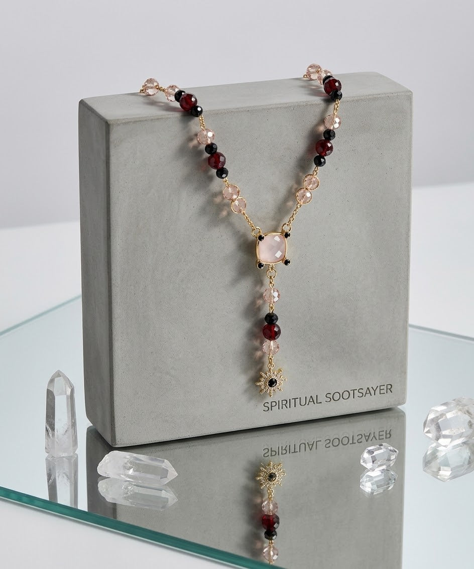 A beaded necklace with a central pale pink stone and dangling gold-toned charms, displayed draped over a gray box labeled SPIRITUAL SOOTSAYER. The necklace includes pink, black, and clear faceted beads.