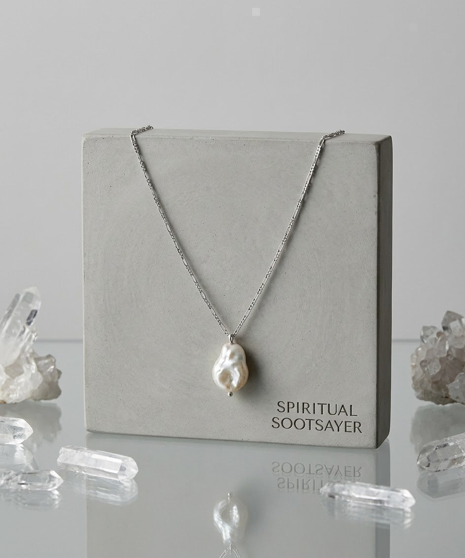 A silver chain necklace with a large, irregular white baroque pearl pendant, displayed on a square gray concrete block labeled 'SPIRITUAL SOOTSAYER'. The display is set on a reflective surface with clear crystal clusters around it against a light gray background.