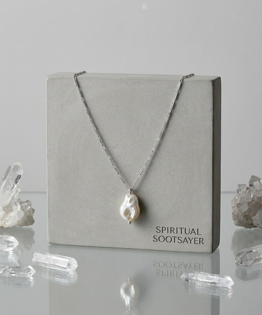 A silver chain necklace with a large, irregular white baroque pearl pendant, displayed on a square gray concrete block labeled 'SPIRITUAL SOOTSAYER'. The display is set on a reflective surface with clear crystal clusters around it against a light gray background.