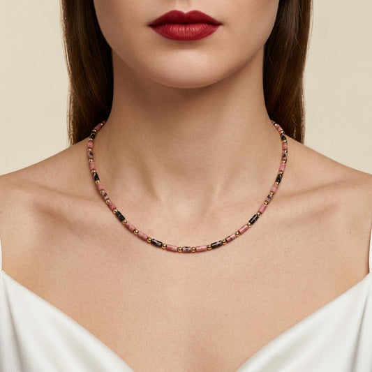 A close-up of a model wearing a delicate beaded necklace with pink, black, and gold beads, styled with a white top against a soft beige background.