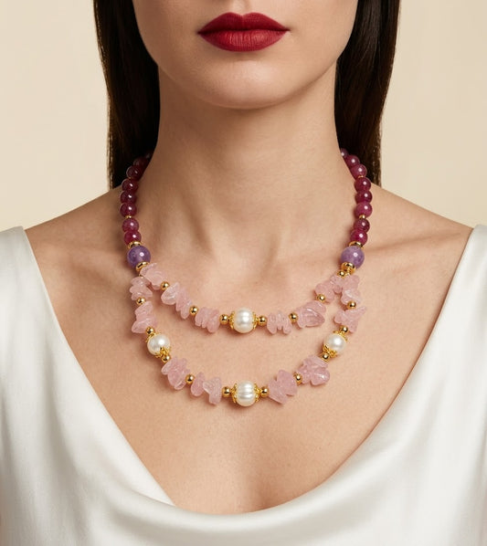 A close-up of a model wearing a double-strand statement necklace featuring pink rose quartz beads, white pearls, and purple gemstone beads with gold spacer rings, paired with a white satin top.