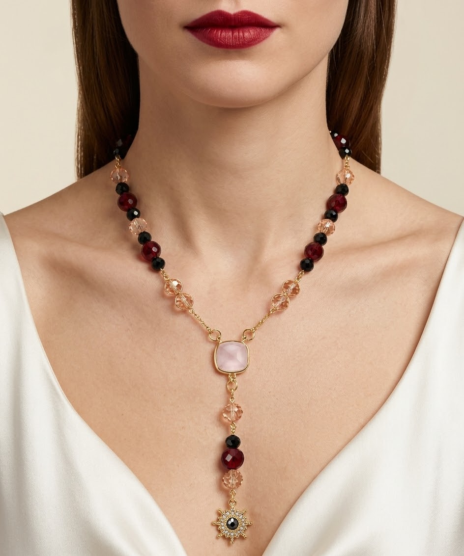 A close-up of a woman wearing a gold-toned V-neck necklace with dark red and black beads, a pale pink stone, and a dangling starburst motif, paired with a white top.