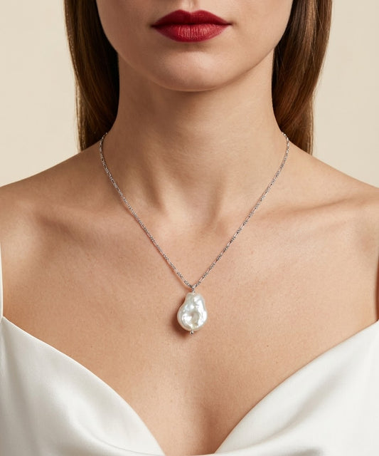 A close-up of a model wearing a delicate silver chain necklace with a large, irregular white baroque pearl pendant, styled with a white satin top against a warm beige background.
