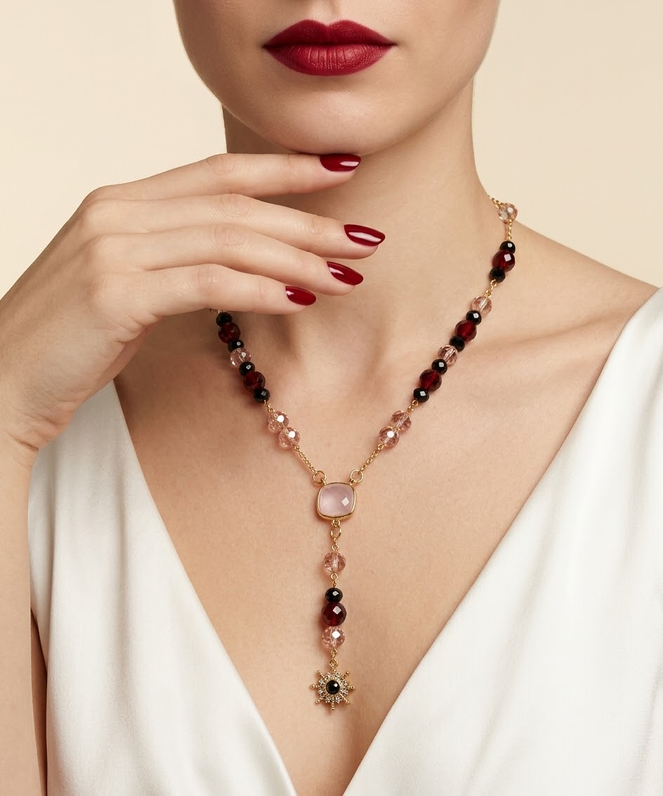 A close-up of a model wearing a delicate gold necklace with dark red, pink, and clear beads, featuring a central pale pink stone and a dangling gold starburst charm.