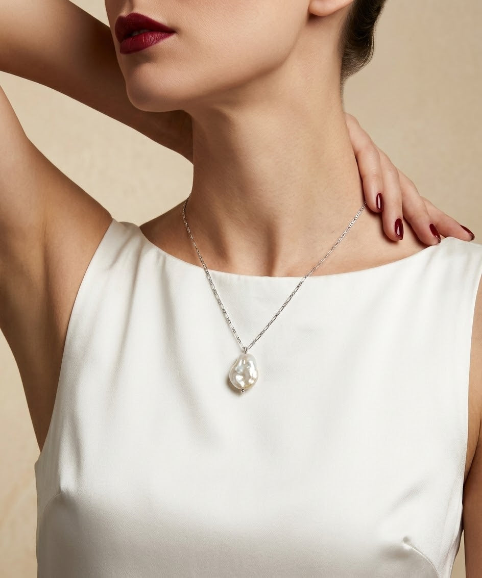 A close-up of a model wearing a delicate silver chain necklace with a teardrop-shaped white pearl pendant, styled with a white sleeveless top against a warm beige background.