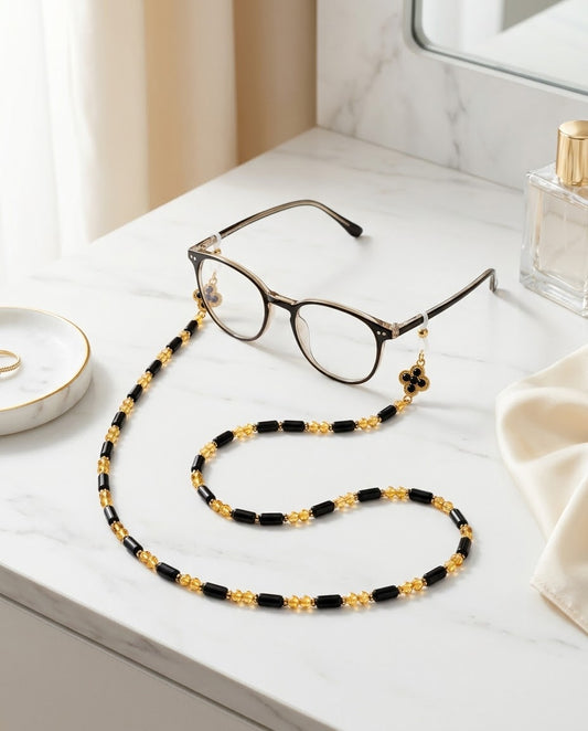 A pair of round metal-framed eyeglasses resting on a white marble surface, attached to a long black-and-gold beaded chain with small clover charms. A perfume bottle, folded cream fabric, and a small dish are visible in a softly lit interior setting.