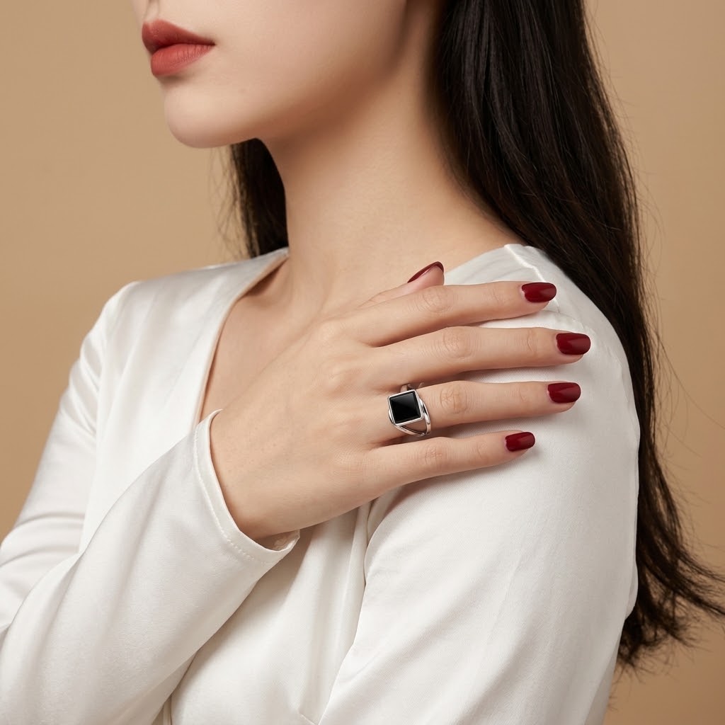 A close-up lifestyle shot of a woman wearing a silver ring with a black rectangular stone on her finger, dressed in a white top against a warm beige background.