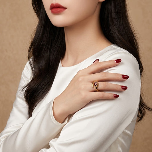 A close-up of a woman wearing a gold ring with a red oval stone on her hand, which is resting on her shoulder. She is wearing a white satin long-sleeve top against a warm beige background.
