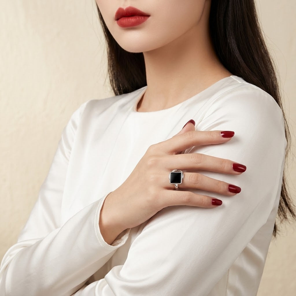 A close-up of a woman wearing a white long-sleeve top, showcasing a silver ring with a square black gemstone on her finger. She has dark hair, red lipstick, and red manicured nails, set against a soft beige textured background.