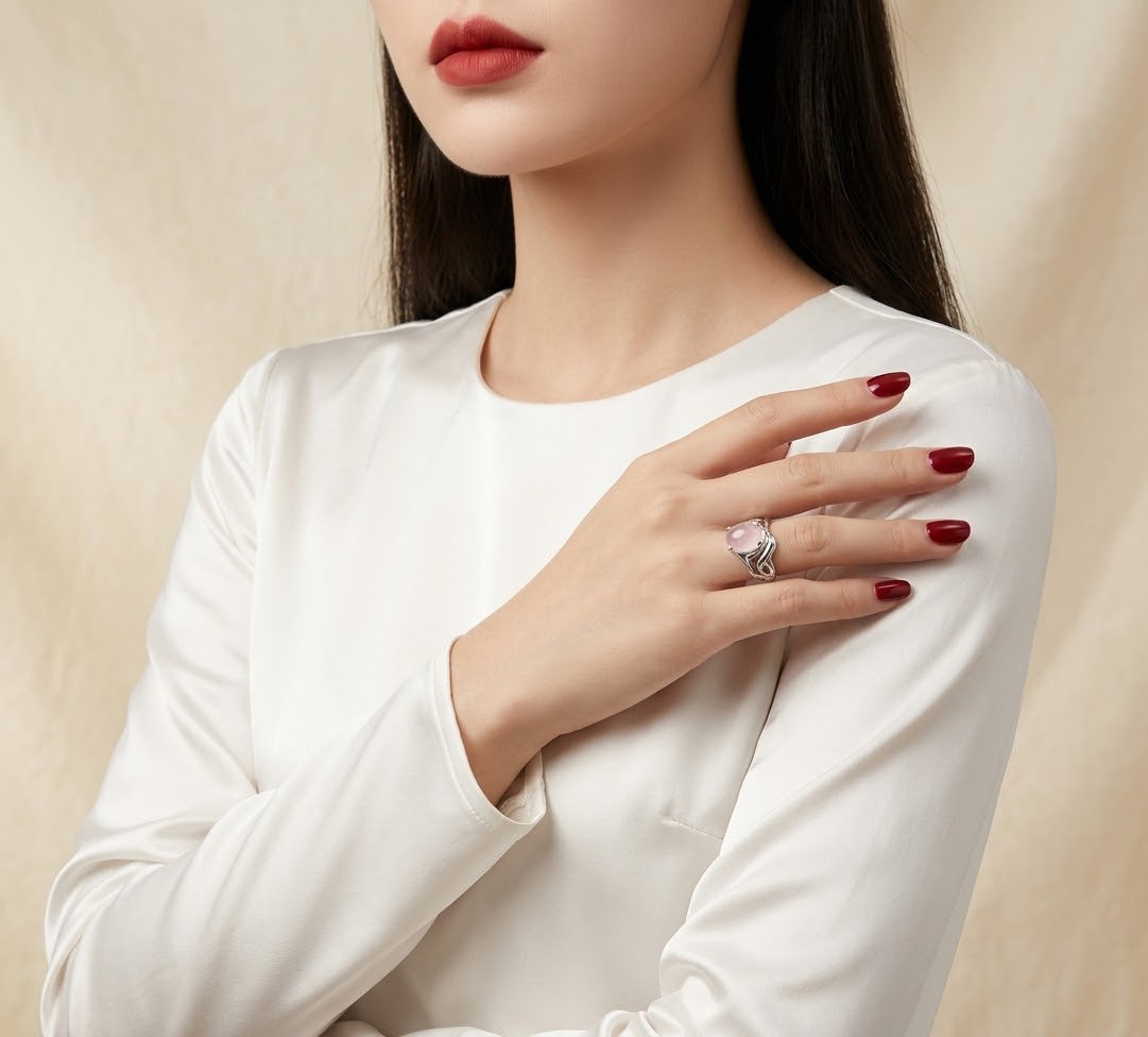 A close-up of a woman wearing a white long-sleeve top, showcasing a silver ring with a pink heart-shaped gemstone on her finger against a soft beige background.