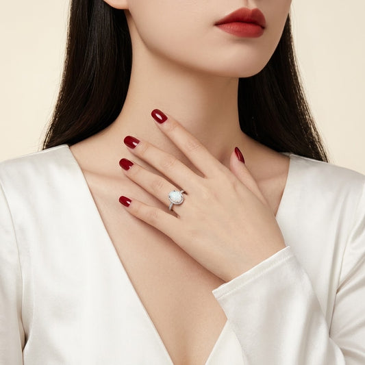 A close-up of a woman wearing a silver ring with a round white stone, styled with dark red nail polish and a white satin top against a soft beige background.