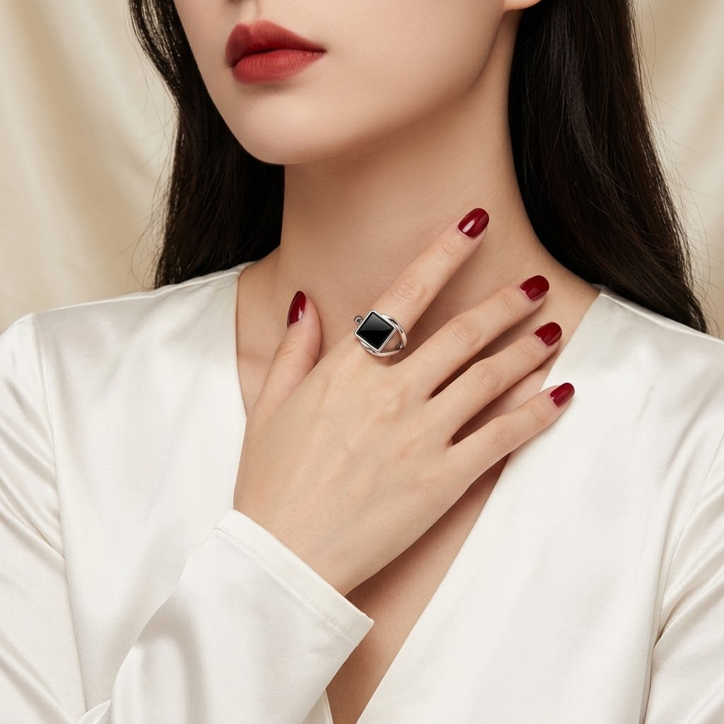 A close-up of a woman wearing a silver-toned ring with a rectangular black inlay on her finger, paired with a white satin top and dark red nail polish.