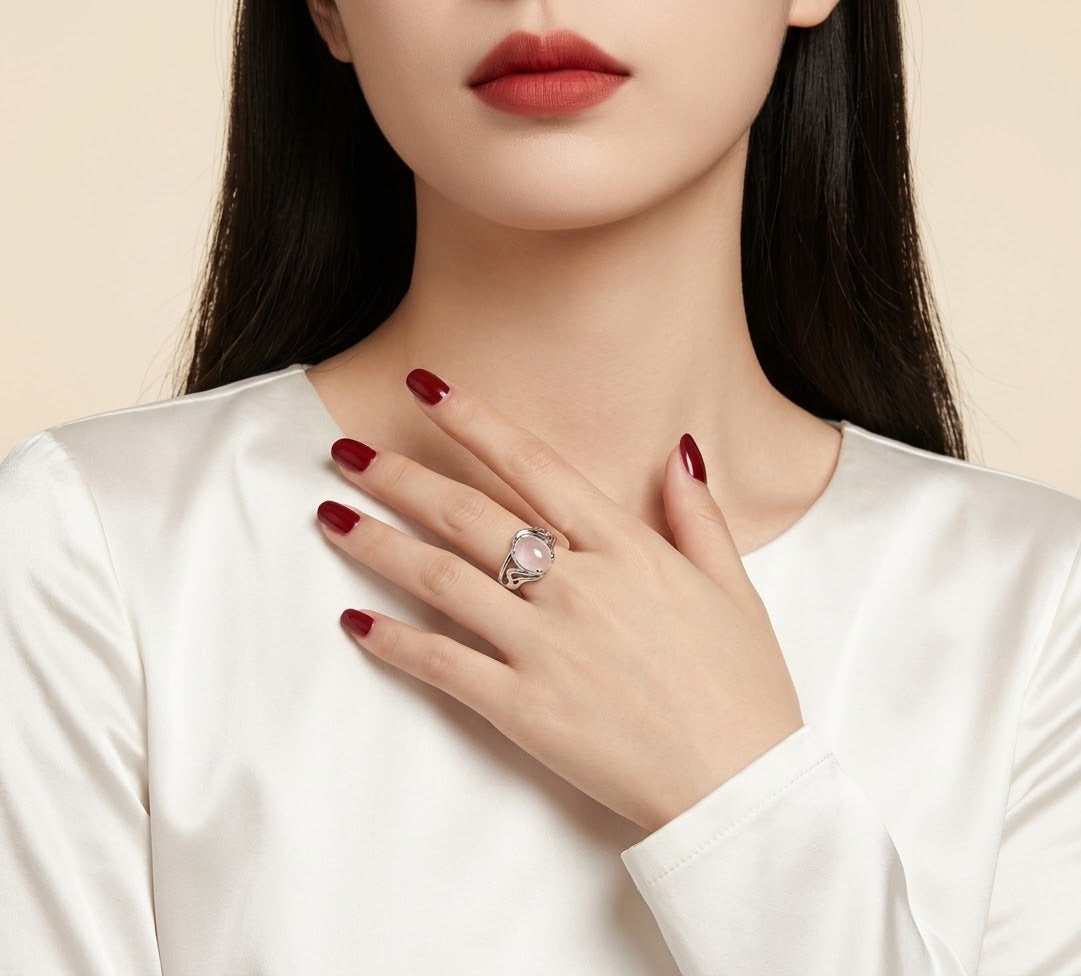 A close-up of a woman wearing a silver ring with a large clear oval gemstone, styled with dark red nail polish and a white satin top against a light beige background.