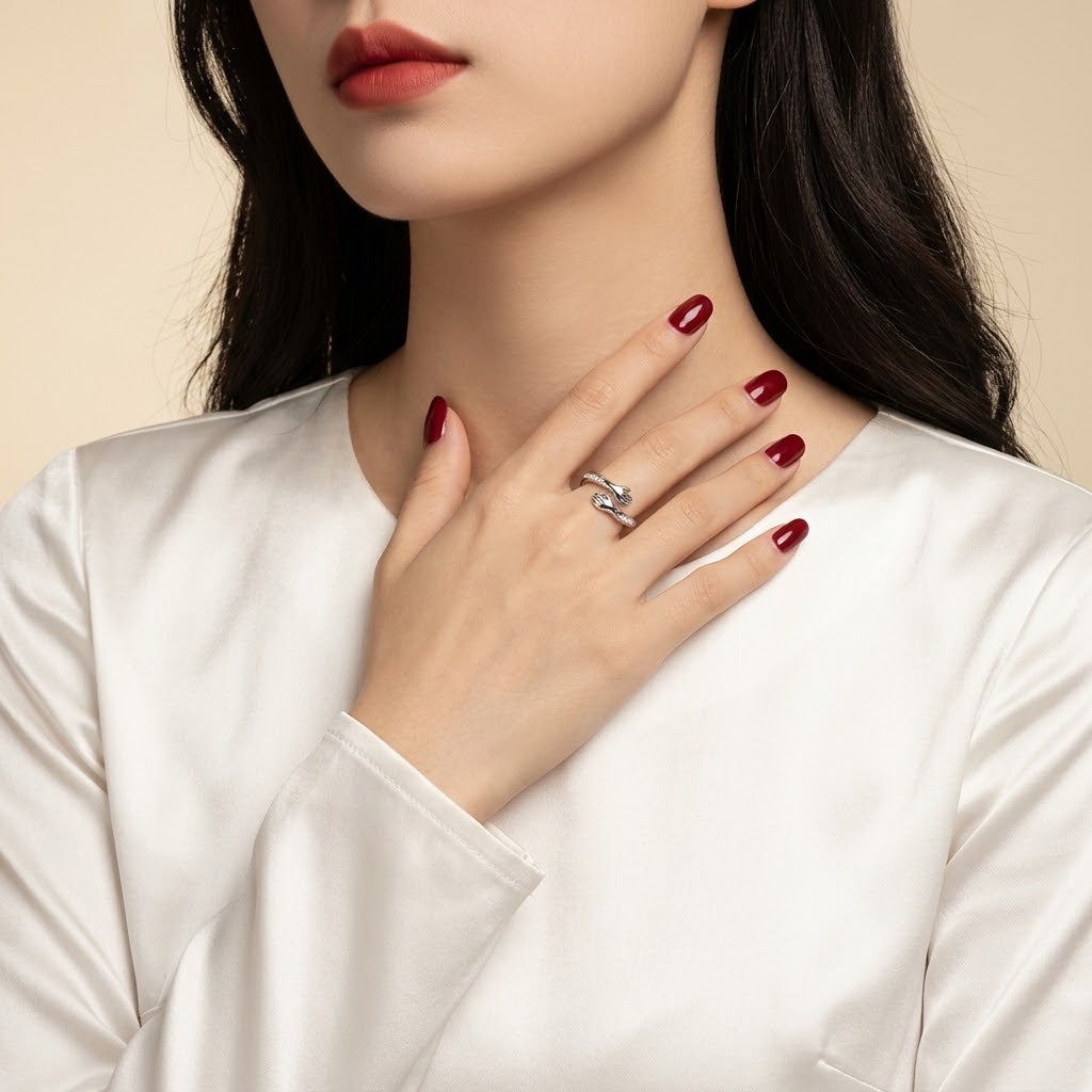 A close-up of a woman wearing a silver ring with clear gemstones on her finger, with her hand resting on her chest. She is wearing a white satin top against a warm beige background.