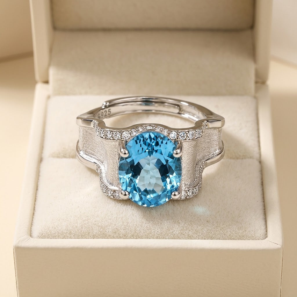 A silver-toned ring with a large oval-cut light blue gemstone and small clear stones along the band, displayed in an open cream jewelry box.