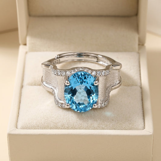 A silver-toned ring with a large oval-cut light blue gemstone and small clear stones along the band, displayed in an open cream jewelry box.