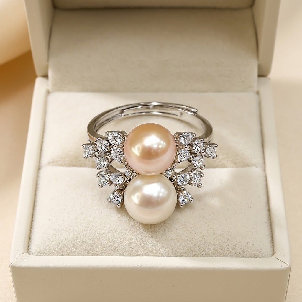A silver-tone ring with two large round pearls, one pale pink and one creamy white, set with small clear stones, displayed in an open cream-colored jewelry box.