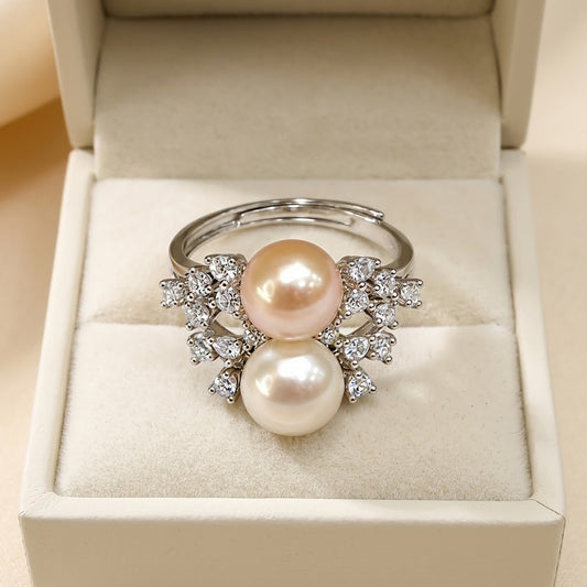 A silver-tone ring with two large round pearls, one pale pink and one creamy white, set with small clear stones, displayed in an open cream-colored jewelry box.