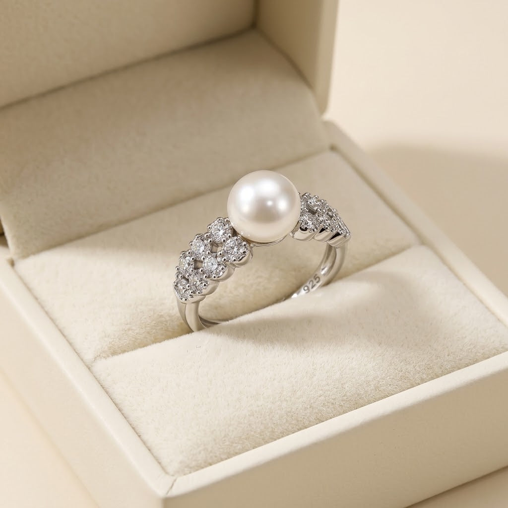 A silver ring with a central white pearl and small clear stones on either side, displayed in an open cream-colored jewelry box.
