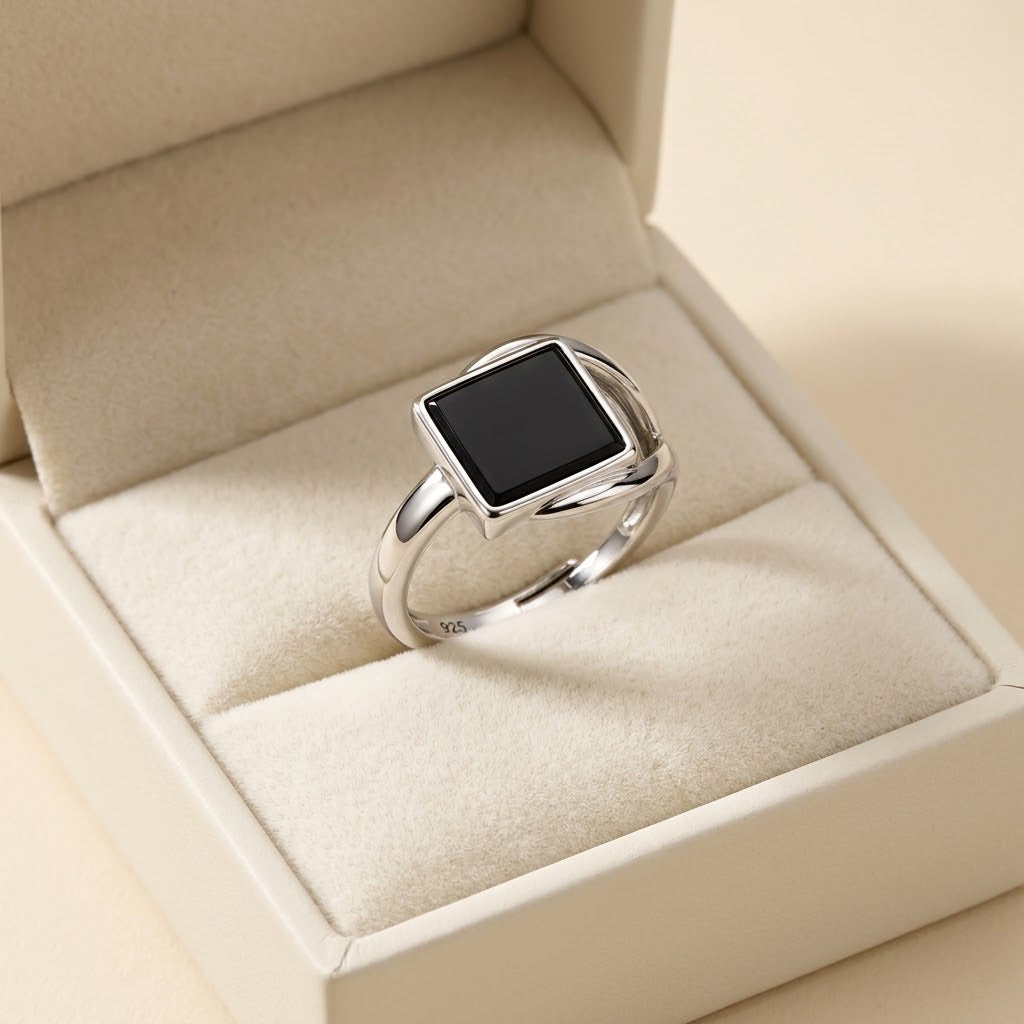 A silver ring with a square black face sits in a cream-colored jewelry box.