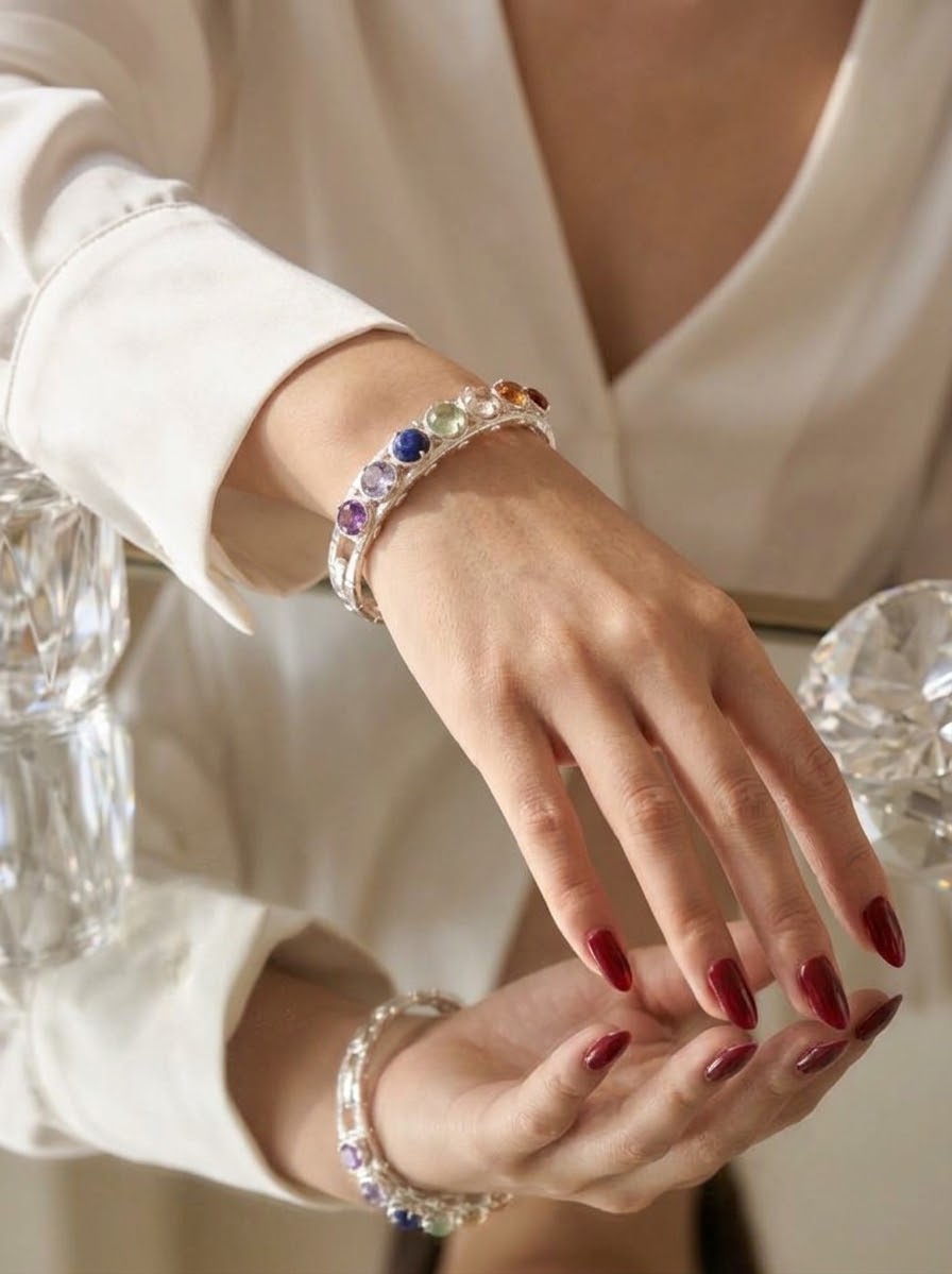 A close-up of a person wearing a silver-toned bracelet adorned with colorful round gemstones, paired with a matching beaded bracelet, set against a soft cream-colored outfit.