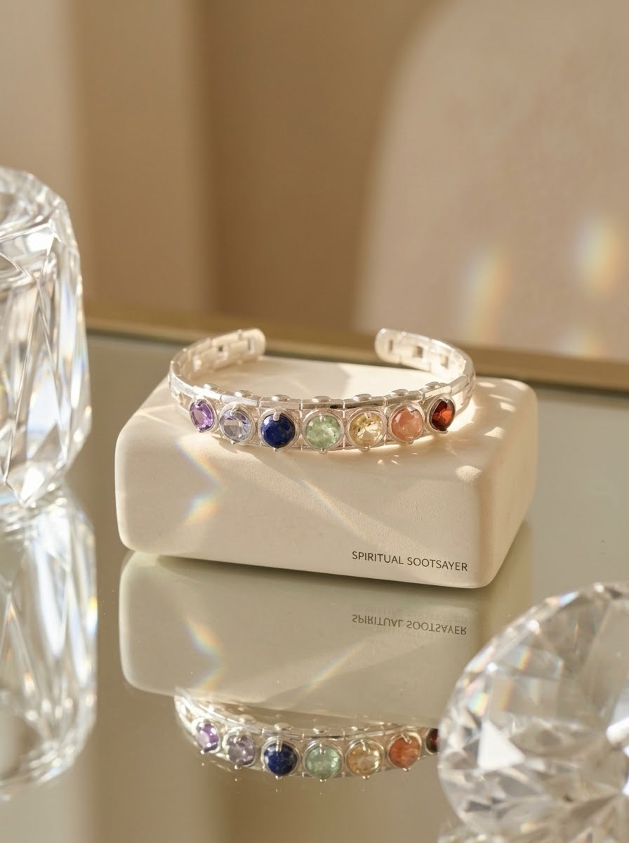 A silver-toned cuff bracelet with a row of colorful gemstones, displayed on a cream-colored box labeled 'SPIRITUAL SOOTSAVER' on a reflective surface.