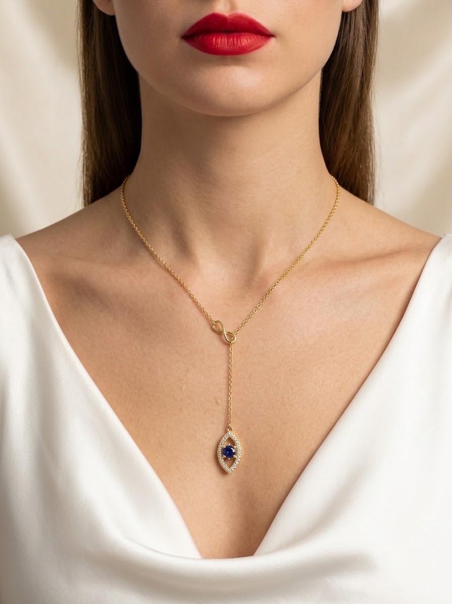 A close-up of a model wearing a delicate gold Y-necklace with a teardrop-shaped pendant featuring a deep blue center stone and small clear accent stones, paired with a white V-neck top.