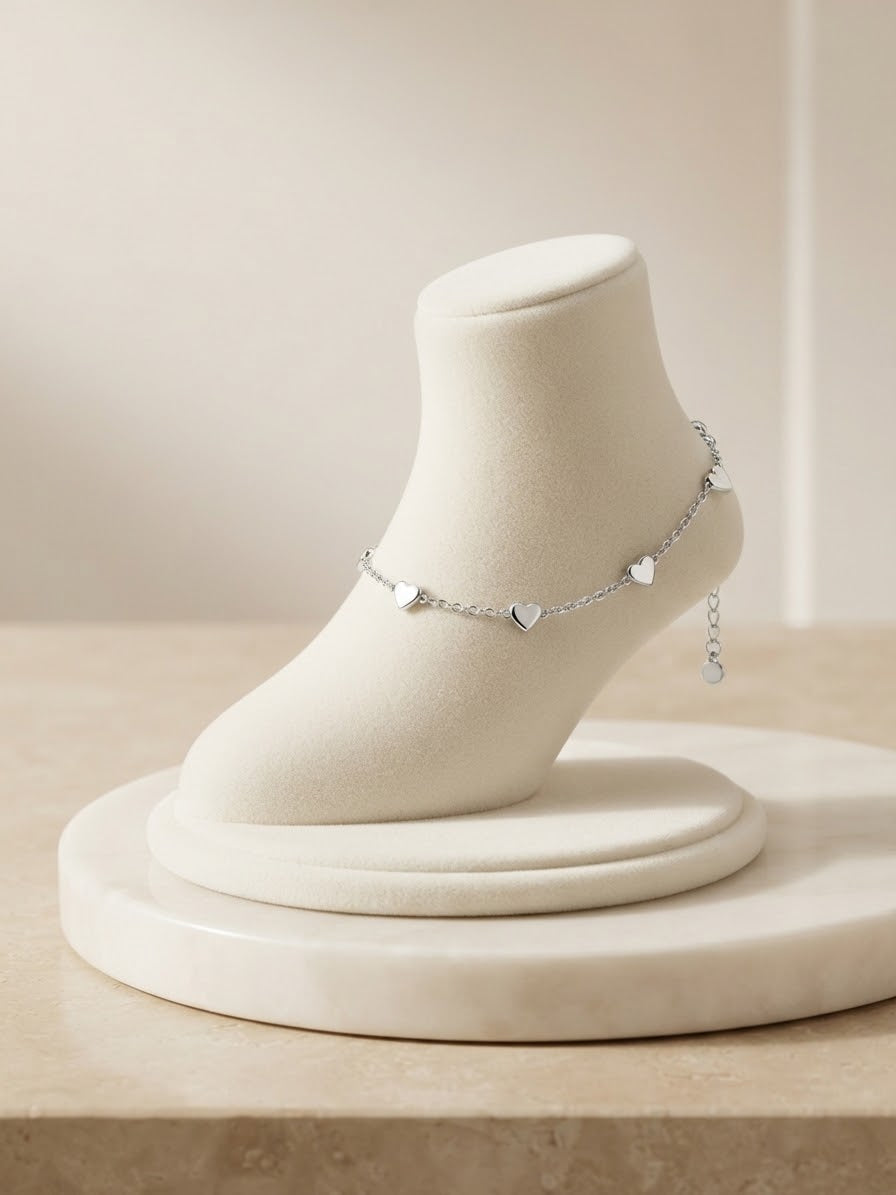 A delicate silver anklet with small heart charms displayed on a cream-colored jewelry bust, set against a soft beige background.