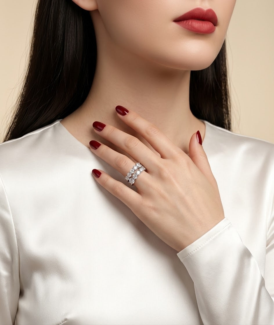 A close-up of a woman wearing a sparkling silver ring with clear stones on her finger, paired with a white satin top and dark red nail polish against a soft beige background.