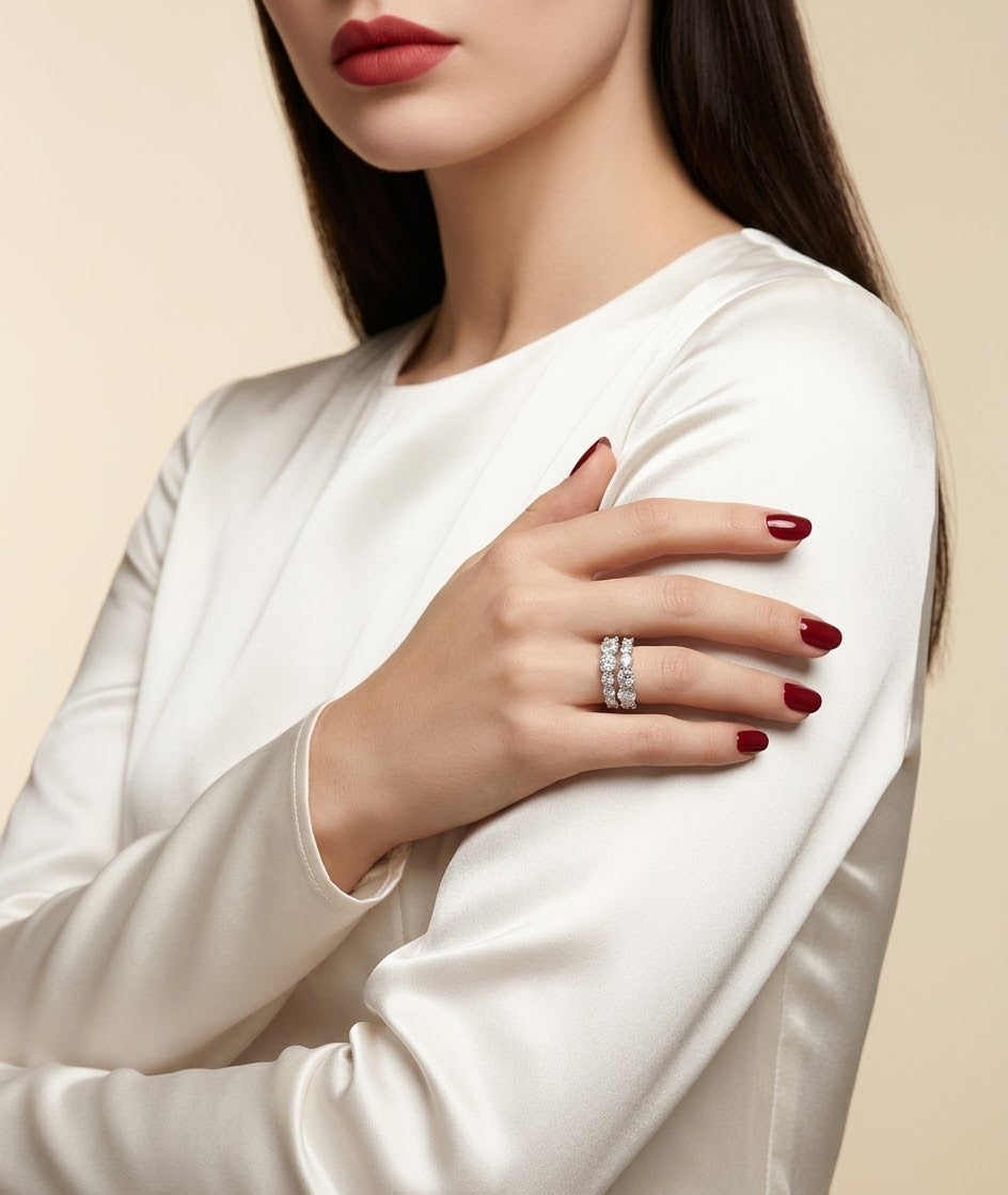 A close-up of a woman wearing a silver ring with clear gemstones on her finger, dressed in a white satin long-sleeve top against a warm beige background.