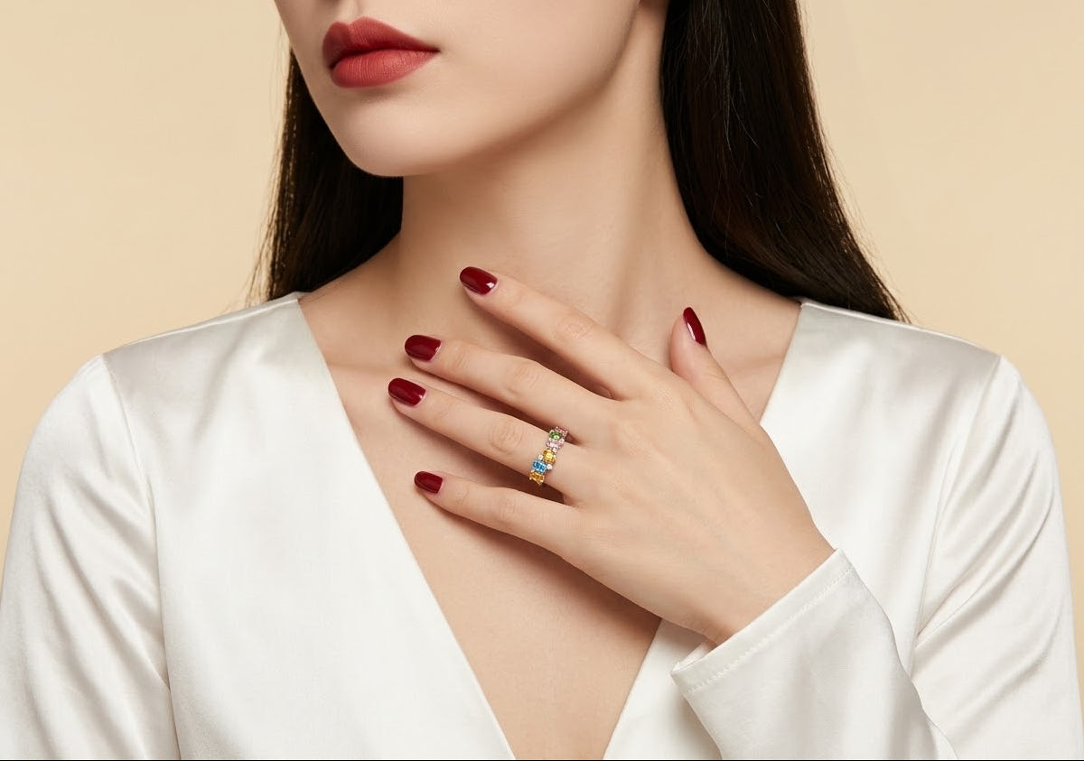 A close-up lifestyle shot of a woman wearing a delicate silver ring with a small blue and red gemstone cluster, styled with dark red nail polish and a white satin top against a warm beige background.