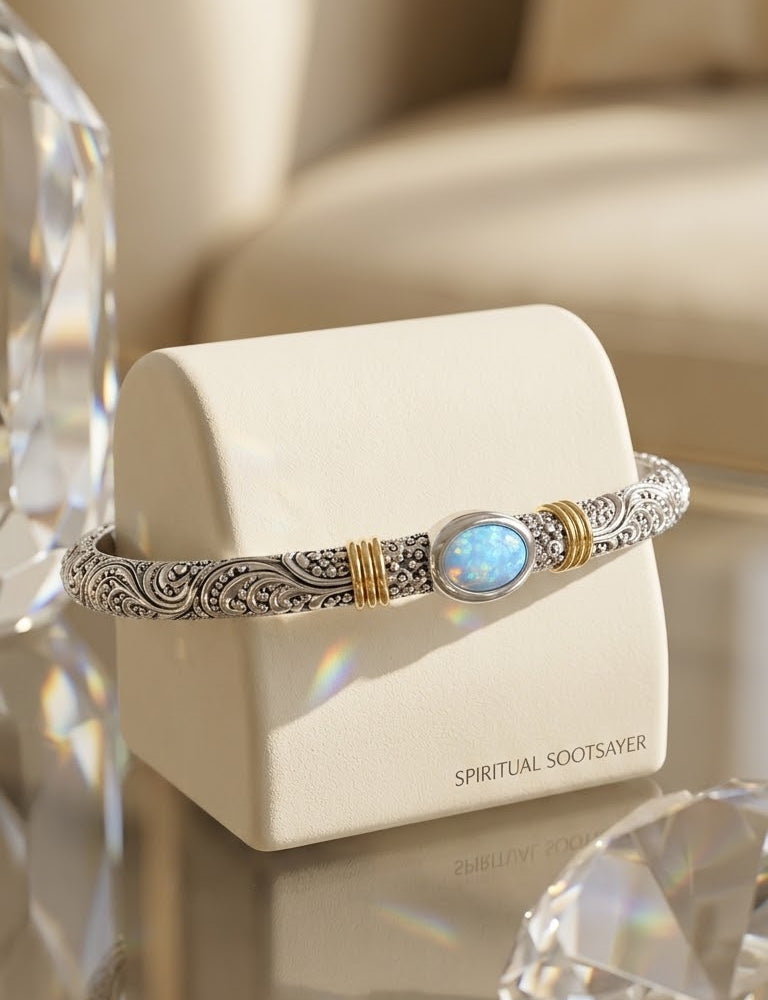 A silver-toned filigree bangle bracelet with gold accents and a round blue stone, displayed on a cream jewelry stand on a glass table with crystal props.