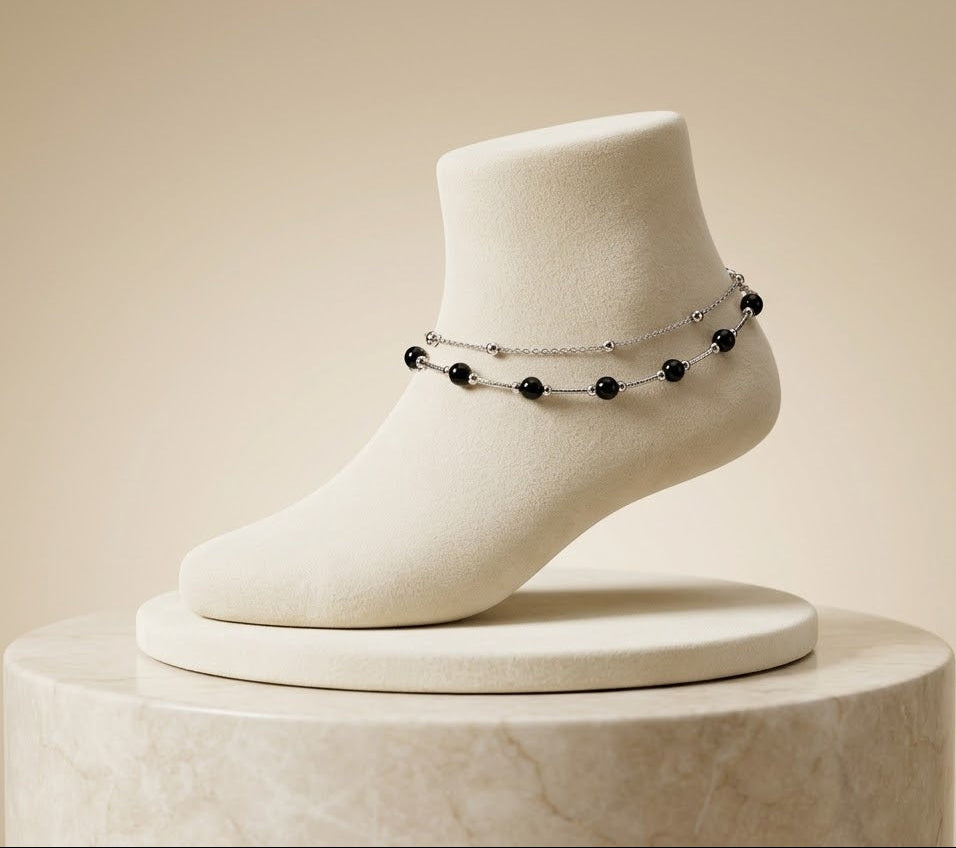 A delicate silver chain anklet with small black beads displayed on a white foot-shaped jewelry stand, set against a warm beige background.