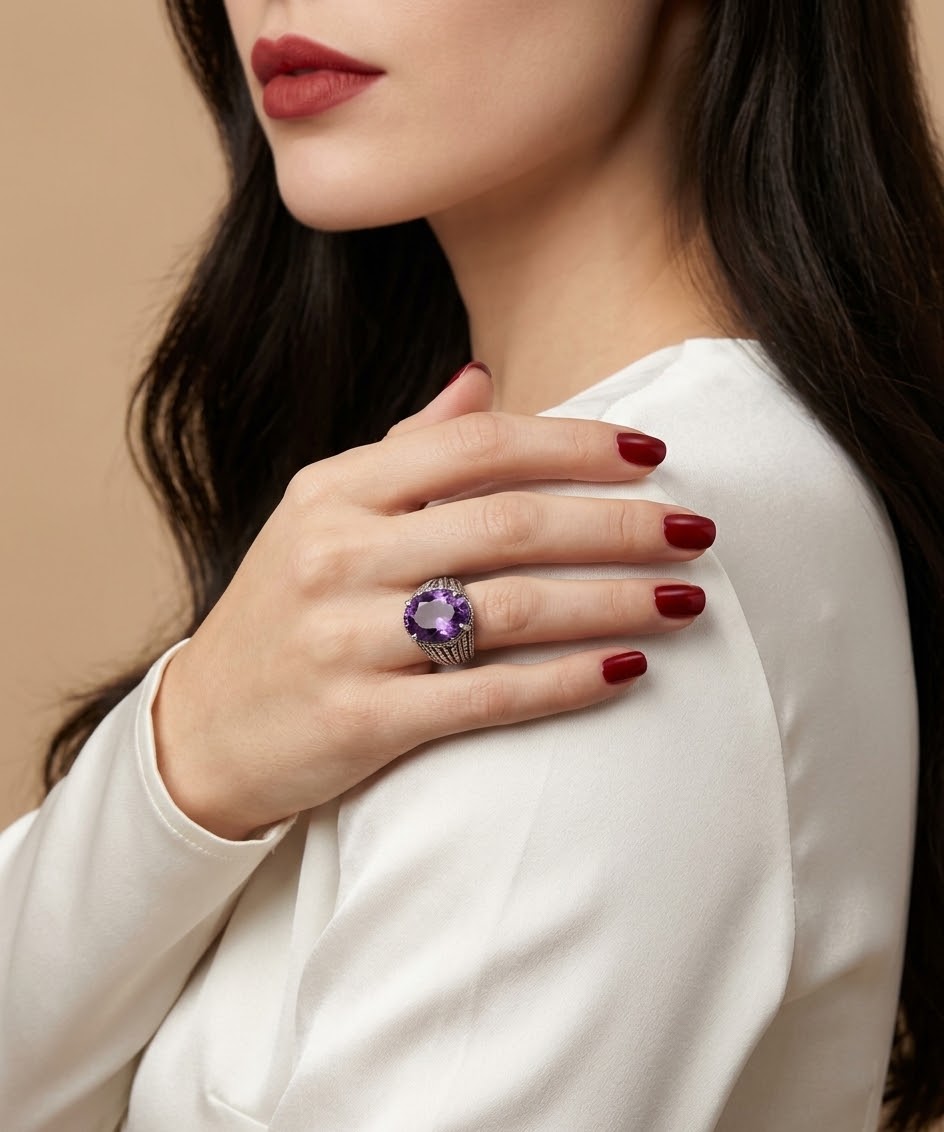 A close-up of a woman wearing a silver ring with a purple oval gemstone, resting on her hand against a warm beige background.