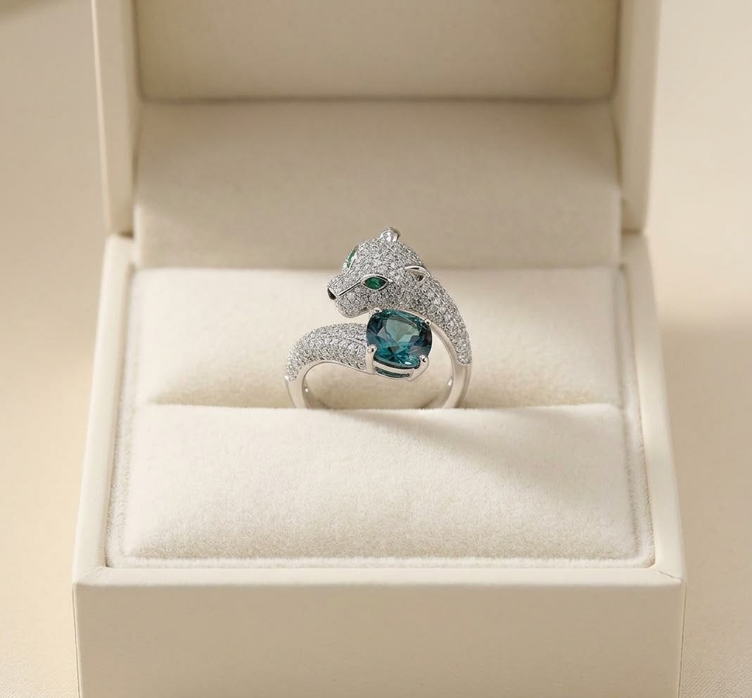 A silver ring with a snake head motif and a central blue gemstone, displayed inside an open cream-colored jewelry box.