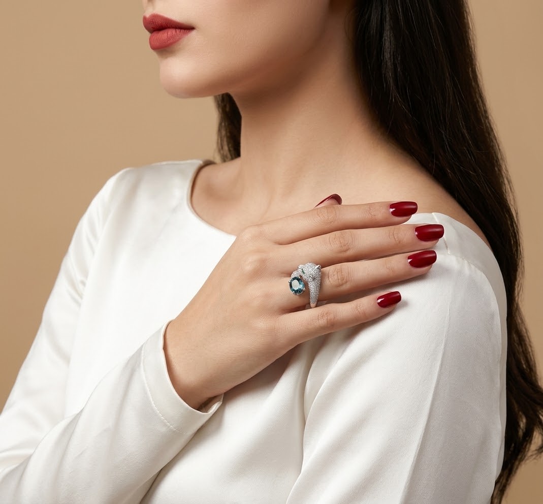 A close-up lifestyle shot of a woman wearing a silver ring with a round blue gemstone, styled with a white long-sleeve top and dark red nail polish against a solid beige background.