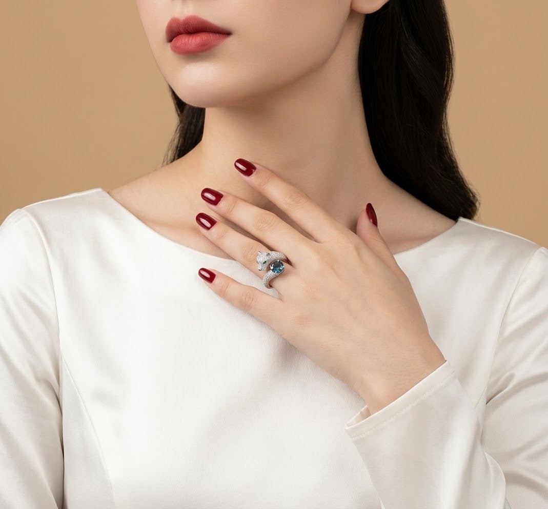 A close-up of a woman wearing a silver ring with a blue gemstone, resting her hand on her chest. She is wearing a white satin top against a solid beige background.