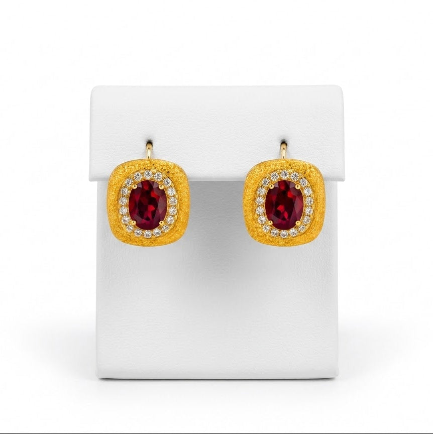 A pair of gold square stud earrings with oval red gemstones and small clear stones in a halo setting, displayed on a white rectangular stand against a white background.