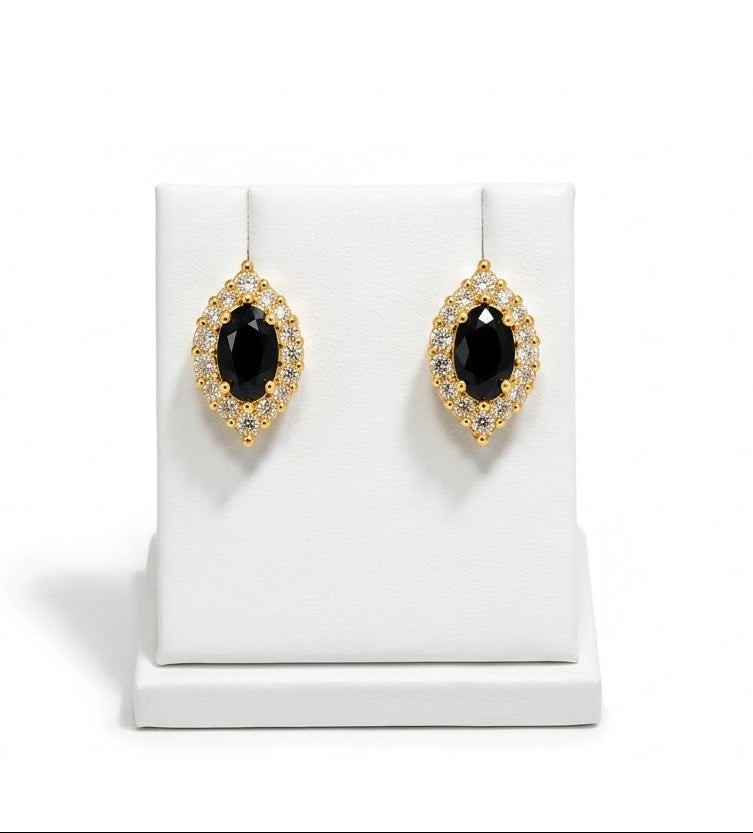 A pair of gold teardrop-shaped drop earrings with oval black centers and small clear stones around the edges, displayed on a white jewelry stand against a white background.