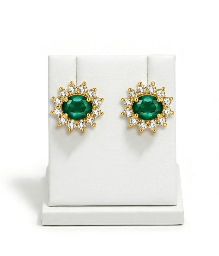 A pair of gold stud earrings with oval green gemstones surrounded by small clear stones, displayed on a white jewelry stand against a white background.
