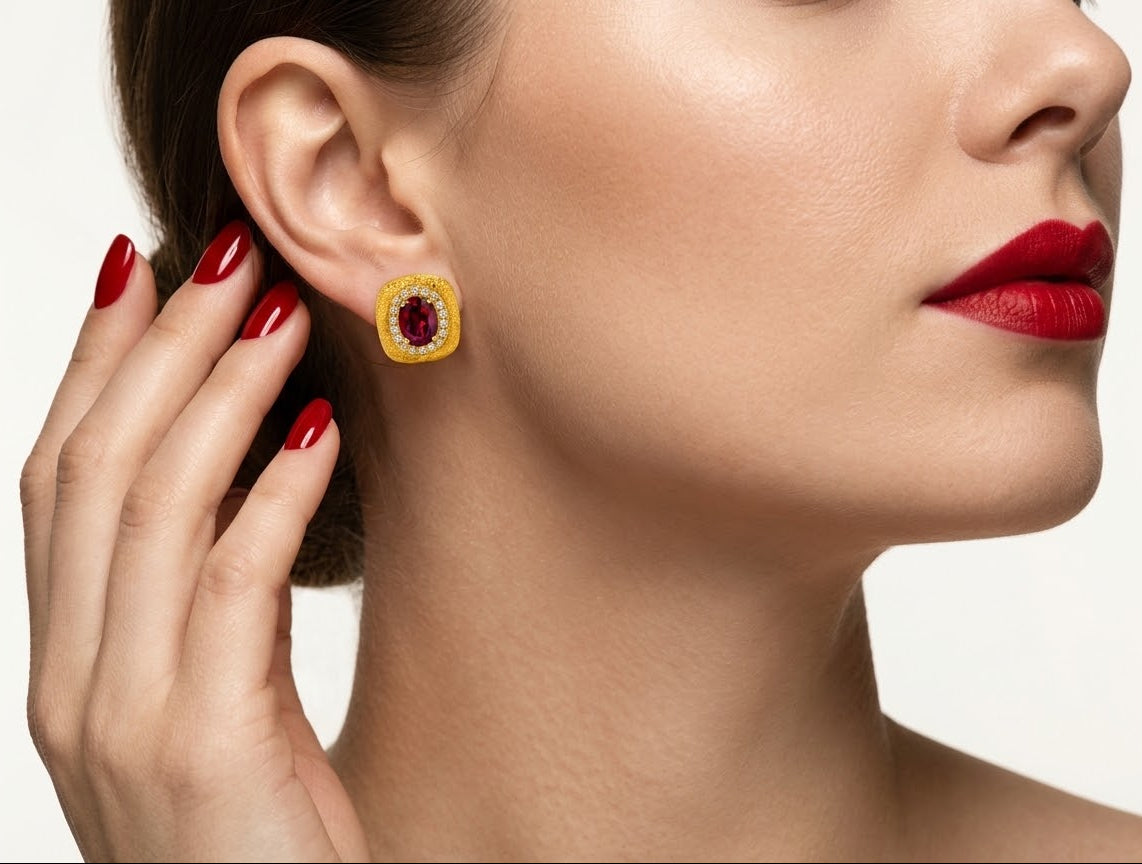 A close-up of a woman wearing a gold square stud earring with a red center stone and a halo of small clear stones, with her hand near her ear against a light gray background.