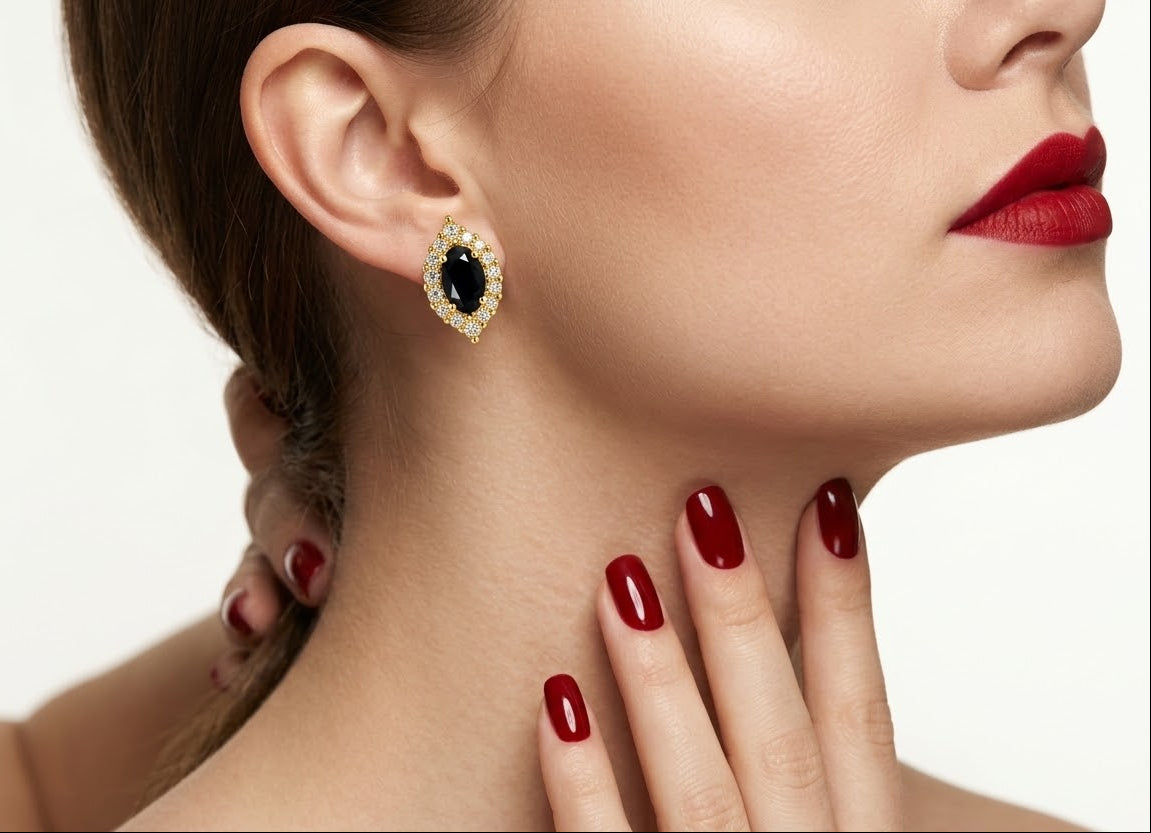 A close-up lifestyle shot of a model wearing a teardrop-shaped gold earring with a black oval center stone and small clear accent stones. The model has dark brown hair, red lipstick, and red manicured nails, posed against a clean white background.